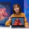 A woman in glasses and a yellow sweater sits at a desk, smiling and holding a laptop displaying digital art of a woman’s face. Behind her is a similar portrait painting. The Photoshop logo appears in the corner, hinting at creative courses online.