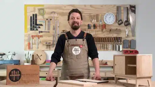 A man wearing an apron stands in a woodworking shop, smiling at the camera. Tools hang neatly on the wall behind him, and a small wooden cabinet and a wooden box labeled MADERÍSTICA hint at courses online for aspiring woodworkers.