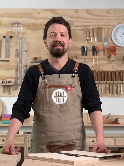 A man wearing an apron stands in a woodworking shop, smiling at the camera. Tools hang neatly on the wall behind him, and a small wooden cabinet and a wooden box labeled MADERÍSTICA hint at courses online for aspiring woodworkers.
