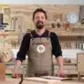 A man wearing an apron stands in a woodworking shop, smiling at the camera. Tools hang neatly on the wall behind him, and a small wooden cabinet and a wooden box labeled MADERÍSTICA hint at courses online for aspiring woodworkers.