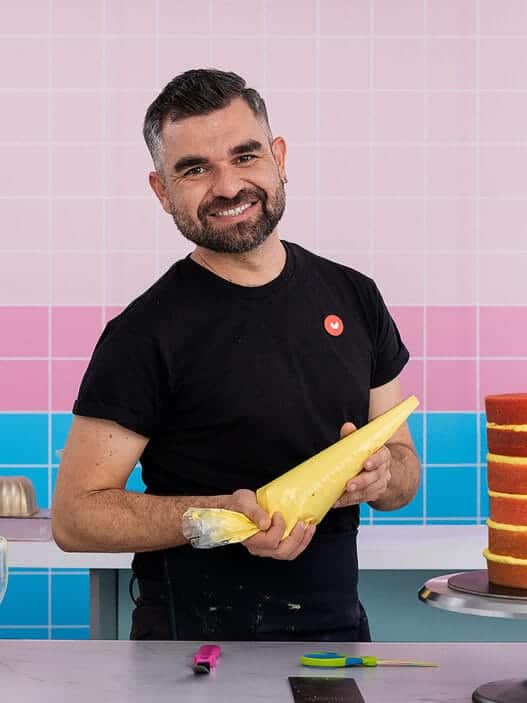 A smiling man stands in a colorful kitchen, holding a piping bag of yellow frosting beside a layered cake. A mixer, utensils, and appliances are on the counter, with a pink neon hello sign—perfect for filming courses online—in the background.