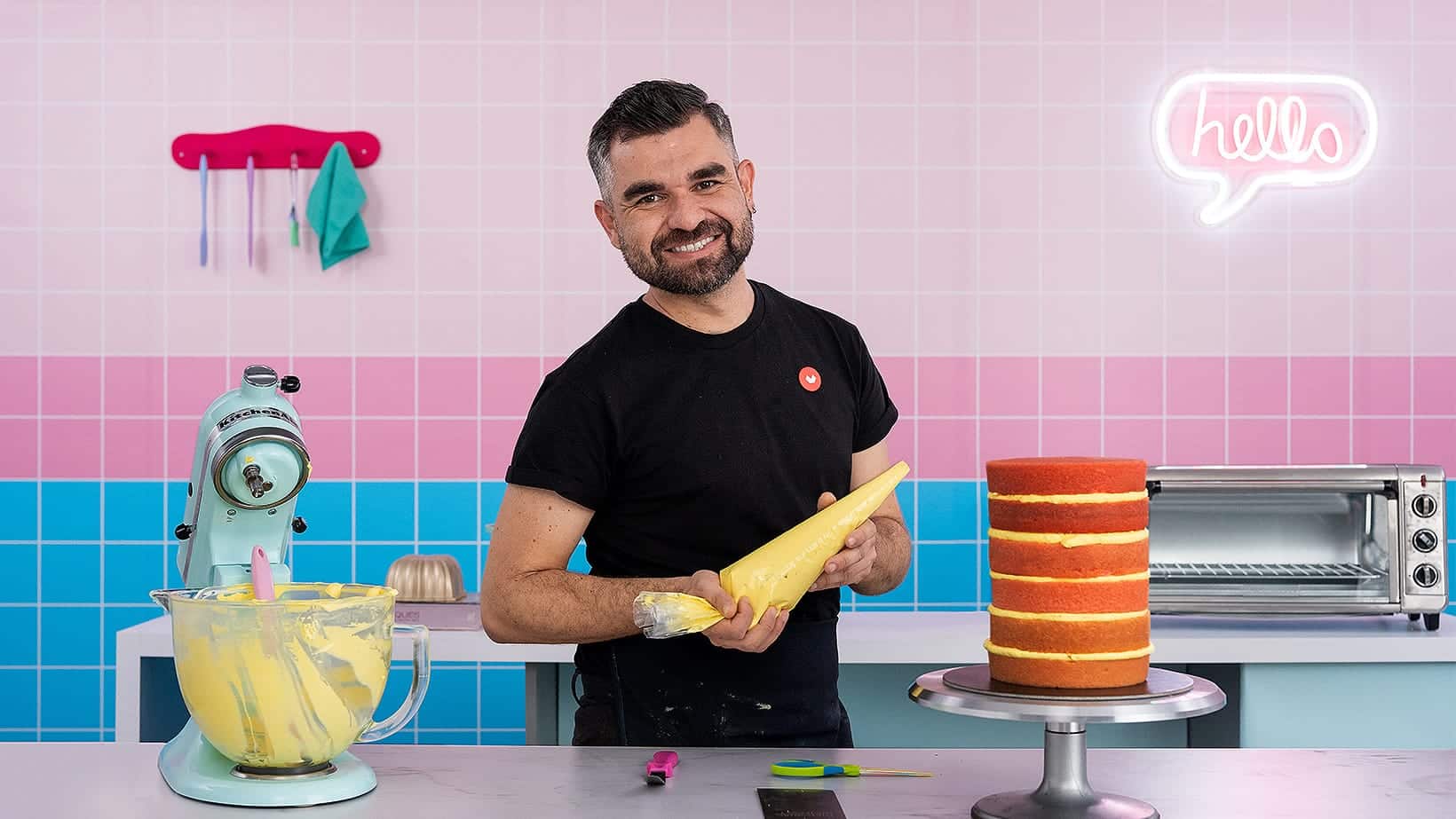 A smiling man stands in a colorful kitchen, holding a piping bag of yellow frosting beside a layered cake. A mixer, utensils, and appliances are on the counter, with a pink neon hello sign—perfect for filming courses online—in the background.