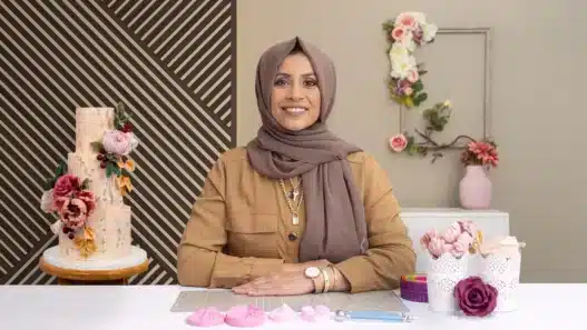 A woman in a brown hijab and tan shirt sits at a table with cake-decorating tools, smiling at the camera. Behind her, floral cakes and flowers are displayed against a wall with geometric and floral decor—perfect inspiration for courses online.