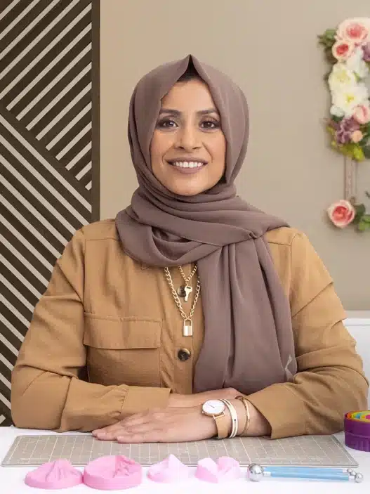 A woman in a brown hijab and tan shirt sits at a table with cake-decorating tools, smiling at the camera. Behind her, floral cakes and flowers are displayed against a wall with geometric and floral decor—perfect inspiration for courses online.