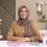 A woman in a brown hijab and tan shirt sits at a table with cake-decorating tools, smiling at the camera. Behind her, floral cakes and flowers are displayed against a wall with geometric and floral decor—perfect inspiration for courses online.