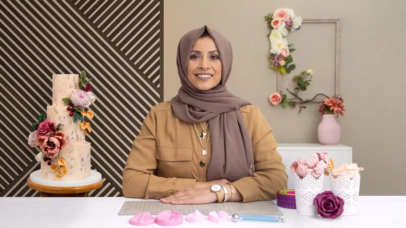 A woman in a brown hijab and tan shirt sits at a table with cake-decorating tools, smiling at the camera. Behind her, floral cakes and flowers are displayed against a wall with geometric and floral decor—perfect inspiration for courses online.