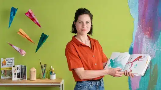 A woman in a red shirt stands in front of a green wall, holding an open picture book. Colorful paper airplanes, art supplies, and books surround her—a vibrant setting perfect for sharing stories or teaching courses online. A painted mural is partially visible on the right.