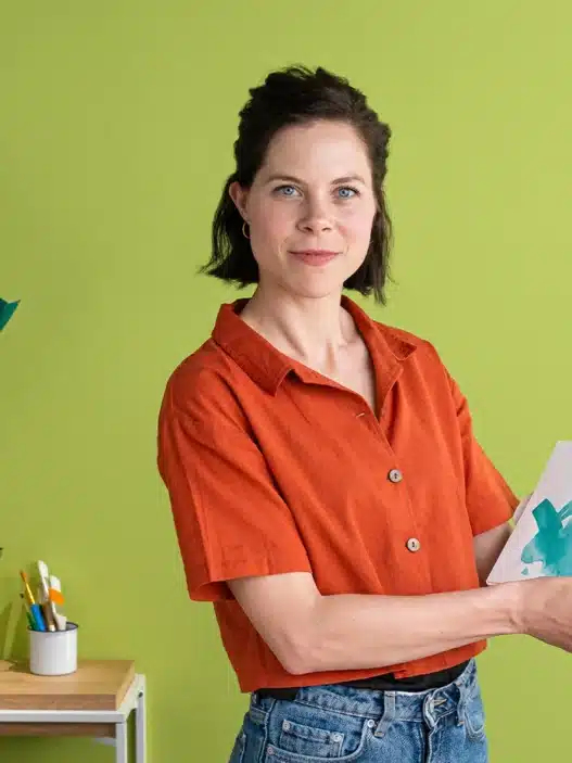 A woman in a red shirt stands in front of a green wall, holding an open picture book. Colorful paper airplanes, art supplies, and books surround her—a vibrant setting perfect for sharing stories or teaching courses online. A painted mural is partially visible on the right.