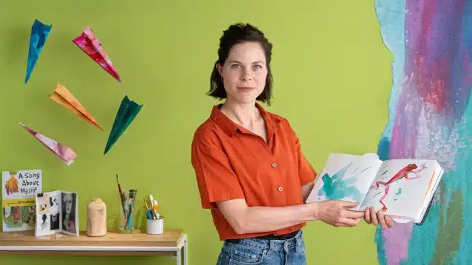 A woman in a red shirt stands in front of a green wall, holding an open picture book. Colorful paper airplanes, art supplies, and books surround her—a vibrant setting perfect for sharing stories or teaching courses online. A painted mural is partially visible on the right.