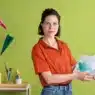 A woman in a red shirt stands in front of a green wall, holding an open picture book. Colorful paper airplanes, art supplies, and books surround her—a vibrant setting perfect for sharing stories or teaching courses online. A painted mural is partially visible on the right.