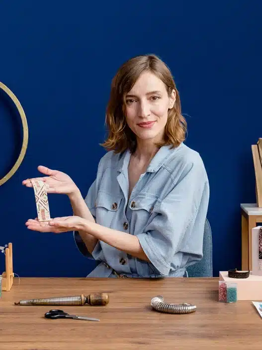A woman sits at a table displaying handmade jewelry and tools, smiling and holding a beaded piece. Behind her, geometric wall designs and shelves with jewelry stands hint at inspiration from courses online.