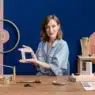 A woman sits at a table displaying handmade jewelry and tools, smiling and holding a beaded piece. Behind her, geometric wall designs and shelves with jewelry stands hint at inspiration from courses online.