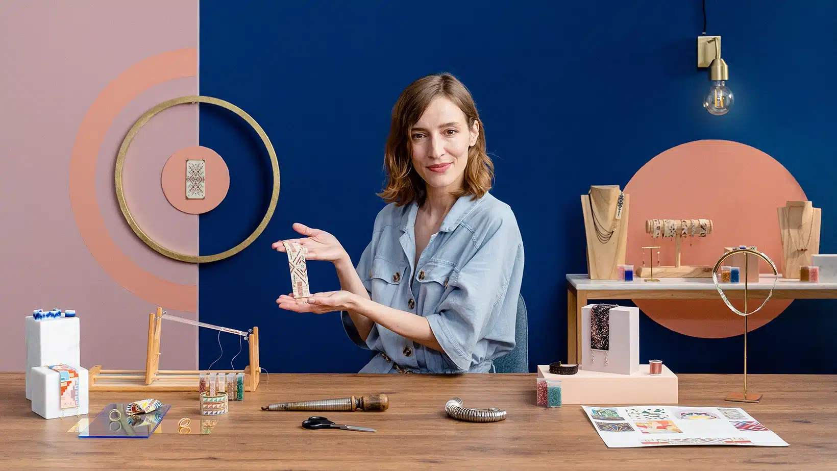 A woman sits at a table displaying handmade jewelry and tools, smiling and holding a beaded piece. Behind her, geometric wall designs and shelves with jewelry stands hint at inspiration from courses online.