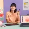 A person with short dark hair sits at a desk holding a stylus, using a drawing tablet and laptop displaying a cute character. Colorful toys and art supplies surround them as they take courses online in a bright, cheerful room.
