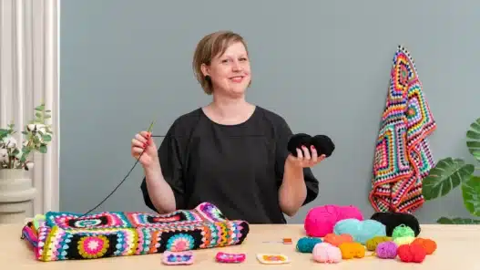 A smiling person sits at a table holding a crochet hook and black yarn, surrounded by colorful yarn balls and crocheted squares. A vibrant crochet blanket drapes in the background, hinting at inspiration for those seeking crochet courses online. A green plant is beside them.