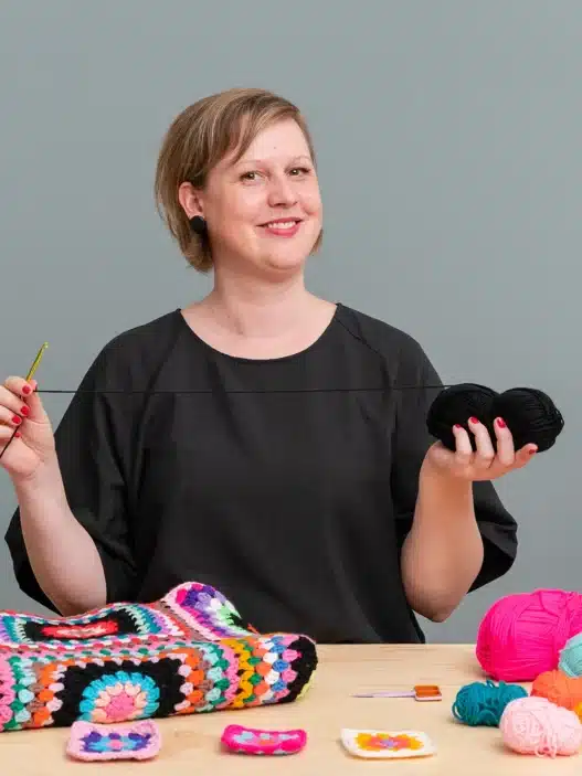 A smiling person sits at a table holding a crochet hook and black yarn, surrounded by colorful yarn balls and crocheted squares. A vibrant crochet blanket drapes in the background, hinting at inspiration for those seeking crochet courses online. A green plant is beside them.