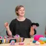 A smiling person sits at a table holding a crochet hook and black yarn, surrounded by colorful yarn balls and crocheted squares. A vibrant crochet blanket drapes in the background, hinting at inspiration for those seeking crochet courses online. A green plant is beside them.