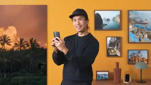 A person in a black cap and sweater smiles while holding a smartphone, standing in front of a yellow wall decorated with framed travel photos, perhaps browsing courses online near a wooden shelf with vases and a small picture.
