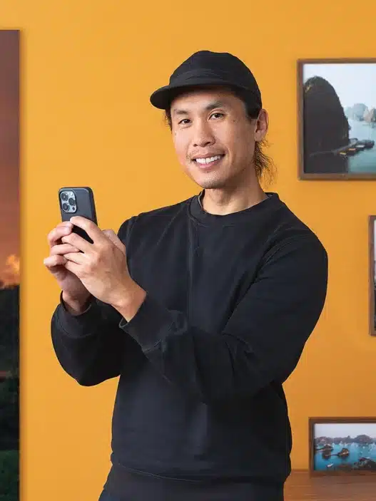 A person in a black cap and sweater smiles while holding a smartphone, standing in front of a yellow wall decorated with framed travel photos, perhaps browsing courses online near a wooden shelf with vases and a small picture.