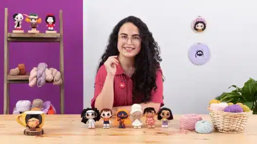 A smiling woman with curly dark hair and glasses sits at a table displaying six handmade dolls. Yarn, a mug, and crochet supplies are on the table—perfect for her courses online. Yarn and framed art hang on the purple and white wall behind her.
