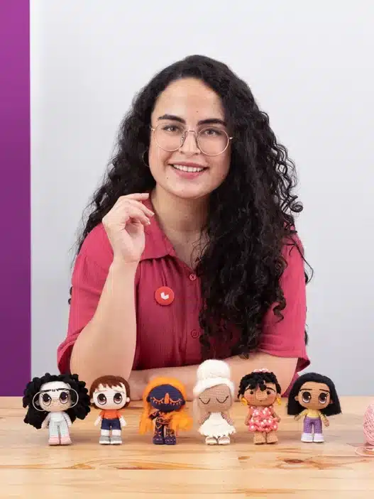 A smiling woman with curly dark hair and glasses sits at a table displaying six handmade dolls. Yarn, a mug, and crochet supplies are on the table—perfect for her courses online. Yarn and framed art hang on the purple and white wall behind her.