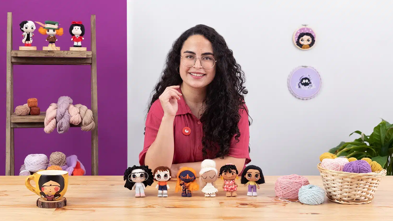 A smiling woman with curly dark hair and glasses sits at a table displaying six handmade dolls. Yarn, a mug, and crochet supplies are on the table—perfect for her courses online. Yarn and framed art hang on the purple and white wall behind her.