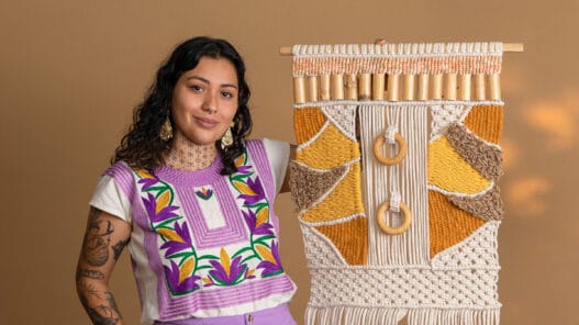 A woman with dark curly hair and tattoos wears a colorful embroidered top and earrings, smiling while holding a large, intricate, yellow and beige macramé wall hanging—showcasing her skills learned from courses online—against a plain tan background.