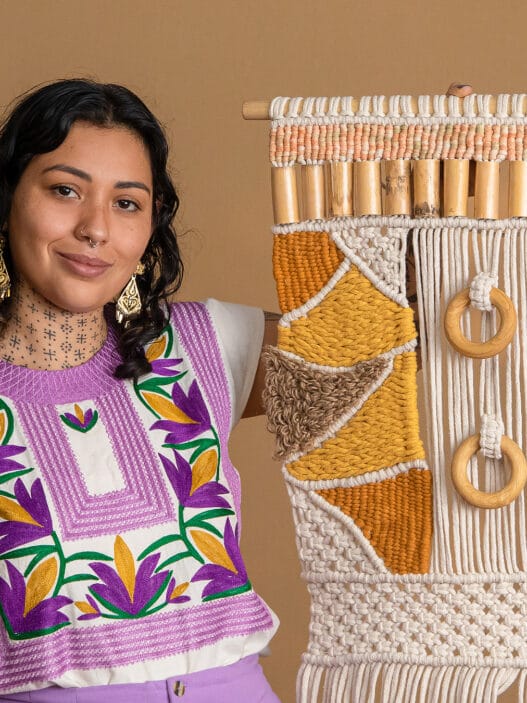 A woman with dark curly hair and tattoos wears a colorful embroidered top and earrings, smiling while holding a large, intricate, yellow and beige macramé wall hanging—showcasing her skills learned from courses online—against a plain tan background.
