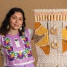 A woman with dark curly hair and tattoos wears a colorful embroidered top and earrings, smiling while holding a large, intricate, yellow and beige macramé wall hanging—showcasing her skills learned from courses online—against a plain tan background.