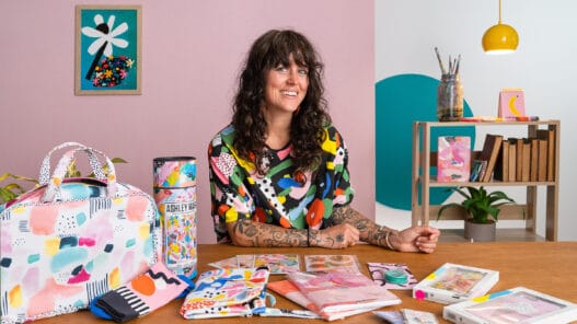 A woman with curly dark hair and tattoos sits at a table covered with colorful, abstract art supplies and products. Smiling in her vibrant shirt, she creates in a lively space filled with framed artwork, bookshelves, and inspiration from courses online.
