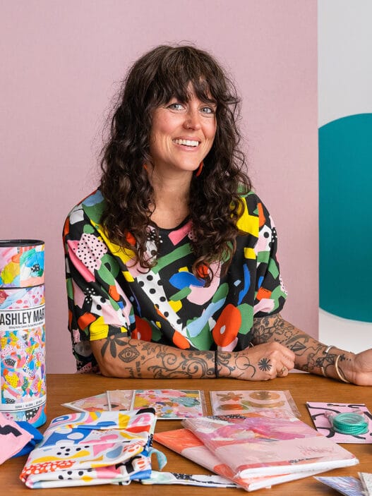 A woman with curly dark hair and tattoos sits at a table covered with colorful, abstract art supplies and products. Smiling in her vibrant shirt, she creates in a lively space filled with framed artwork, bookshelves, and inspiration from courses online.