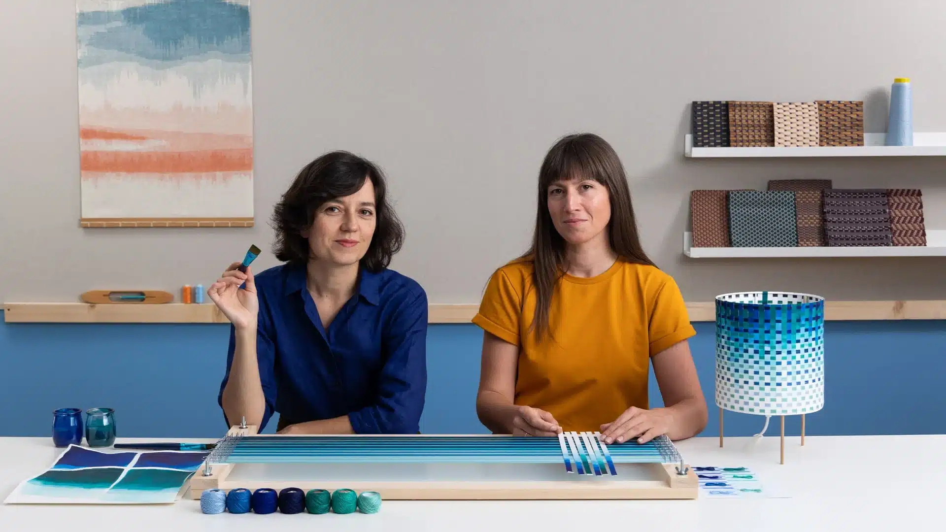 Two women sit at a table with weaving materials and colorful yarns; one discusses courses online as the other holds a tool, while a finished woven lamp and art supplies are displayed in their modern, creative workspace.