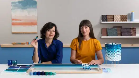 Two women sit at a table with weaving materials and colorful yarns; one discusses courses online as the other holds a tool, while a finished woven lamp and art supplies are displayed in their modern, creative workspace.