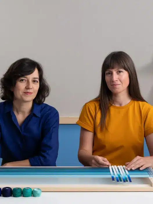 Two women sit at a table with weaving materials and colorful yarns; one discusses courses online as the other holds a tool, while a finished woven lamp and art supplies are displayed in their modern, creative workspace.
