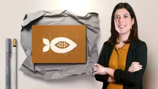 A woman with dark hair, wearing a black jacket and mustard shirt, stands smiling with arms crossed beside a brown card featuring a geometric white seed design on crumpled paper—an inspiring scene for those interested in creative courses online. Two pens are to the left.