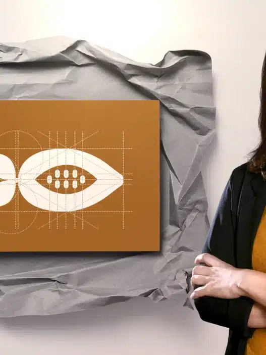 A woman with dark hair, wearing a black jacket and mustard shirt, stands smiling with arms crossed beside a brown card featuring a geometric white seed design on crumpled paper—an inspiring scene for those interested in creative courses online. Two pens are to the left.