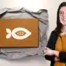 A woman with dark hair, wearing a black jacket and mustard shirt, stands smiling with arms crossed beside a brown card featuring a geometric white seed design on crumpled paper—an inspiring scene for those interested in creative courses online. Two pens are to the left.