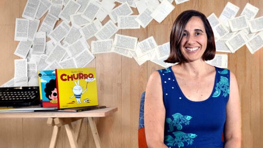 A woman with shoulder-length brown hair sits smiling in front of a wall decorated with scattered book pages. Behind her, a wooden desk holds a typewriter, colorful childrens books, and materials for courses online.