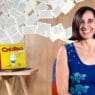 A woman with shoulder-length brown hair sits smiling in front of a wall decorated with scattered book pages. Behind her, a wooden desk holds a typewriter, colorful childrens books, and materials for courses online.