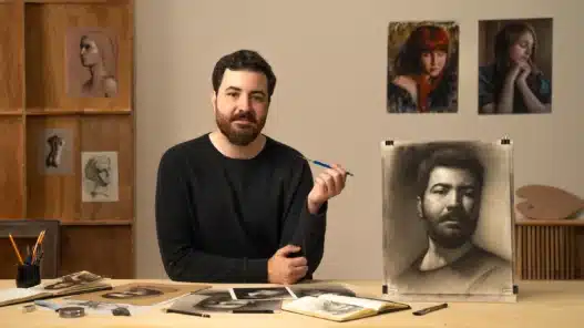 A bearded man sits at a table holding a brush, with a realistic charcoal portrait of himself in front of him. Surrounded by art supplies and sketches, he appears ready to teach courses online, with inspiring artworks hanging on the wall behind him.