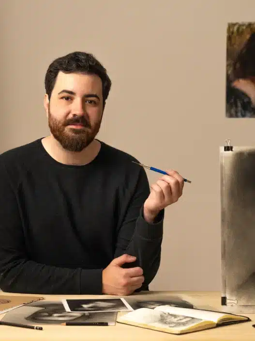 A bearded man sits at a table holding a brush, with a realistic charcoal portrait of himself in front of him. Surrounded by art supplies and sketches, he appears ready to teach courses online, with inspiring artworks hanging on the wall behind him.