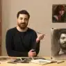 A bearded man sits at a table holding a brush, with a realistic charcoal portrait of himself in front of him. Surrounded by art supplies and sketches, he appears ready to teach courses online, with inspiring artworks hanging on the wall behind him.