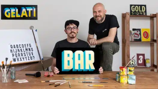Two artists pose in a studio. One sits holding a sign that reads BAR in bold, colorful letters while the other stands beside him. Art supplies and lettered signs fill the space—a creative hub perfect for exploring courses online.