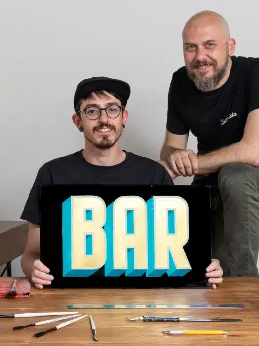 Two artists pose in a studio. One sits holding a sign that reads BAR in bold, colorful letters while the other stands beside him. Art supplies and lettered signs fill the space—a creative hub perfect for exploring courses online.