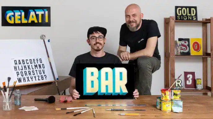 Two artists pose in a studio. One sits holding a sign that reads BAR in bold, colorful letters while the other stands beside him. Art supplies and lettered signs fill the space—a creative hub perfect for exploring courses online.
