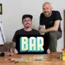 Two artists pose in a studio. One sits holding a sign that reads BAR in bold, colorful letters while the other stands beside him. Art supplies and lettered signs fill the space—a creative hub perfect for exploring courses online.