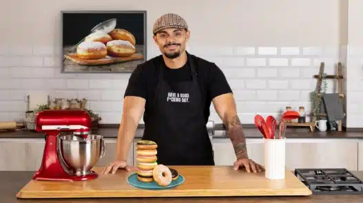 A smiling man in a cap and black apron stands in a modern kitchen behind a stack of donuts on a plate, ready to teach courses online. A red stand mixer, utensils, and donut photo on the wall are also visible.