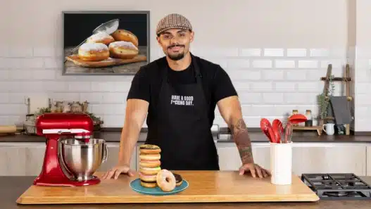 A smiling man in a cap and black apron stands in a modern kitchen behind a stack of donuts on a plate, ready to teach courses online. A red stand mixer, utensils, and donut photo on the wall are also visible.