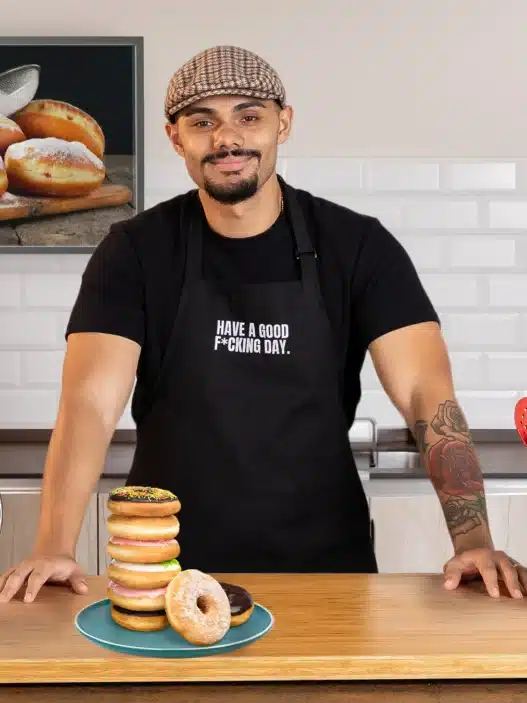 A smiling man in a cap and black apron stands in a modern kitchen behind a stack of donuts on a plate, ready to teach courses online. A red stand mixer, utensils, and donut photo on the wall are also visible.