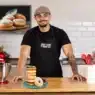 A smiling man in a cap and black apron stands in a modern kitchen behind a stack of donuts on a plate, ready to teach courses online. A red stand mixer, utensils, and donut photo on the wall are also visible.